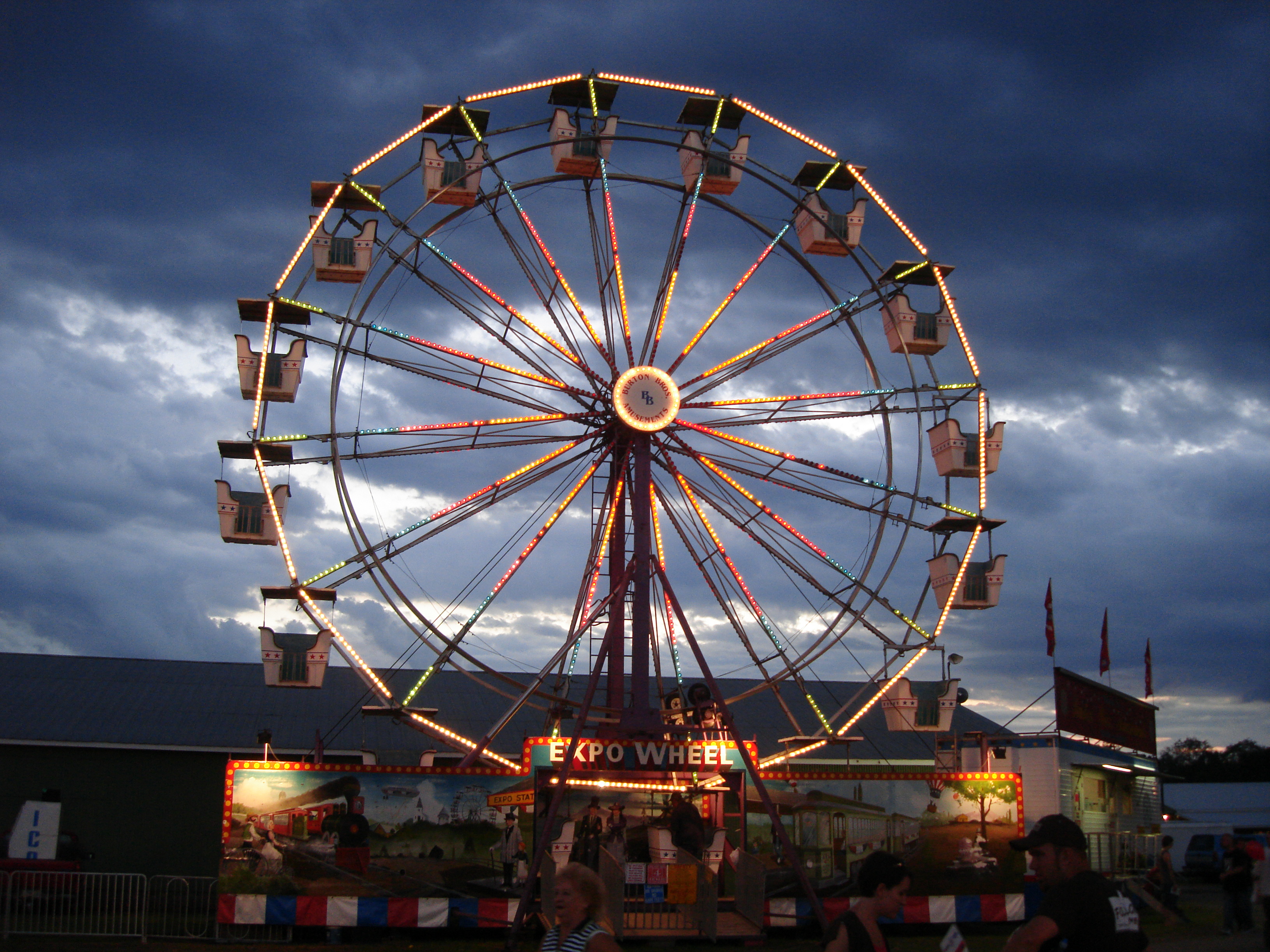Photo of a Ferris Wheel with bright colored lights, surrounded by cloudy skies.