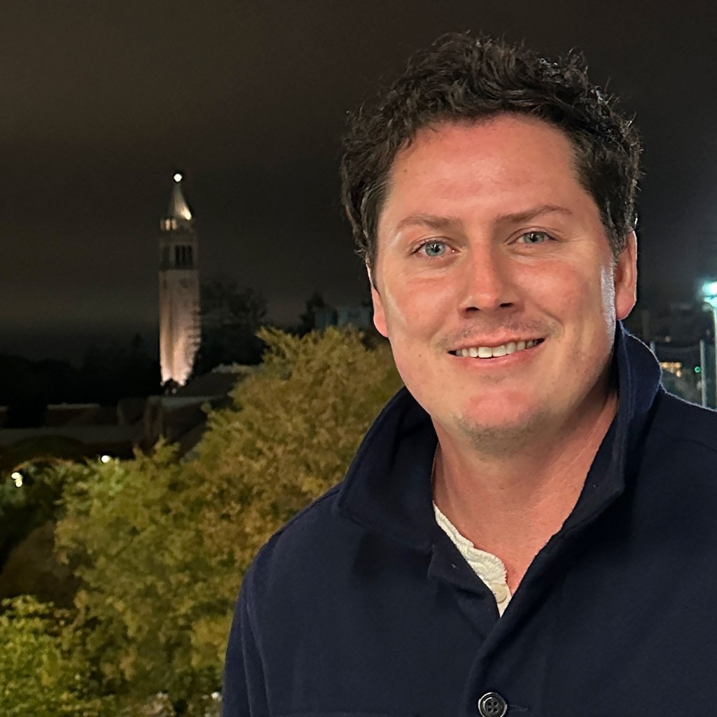 A man smiling at night with buildings and trees in the background, including a tower visible in the distance.