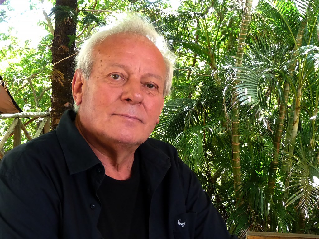 A portrait of a man with grey hair wearing a black shirt, seated outdoors amidst lush green foliage.