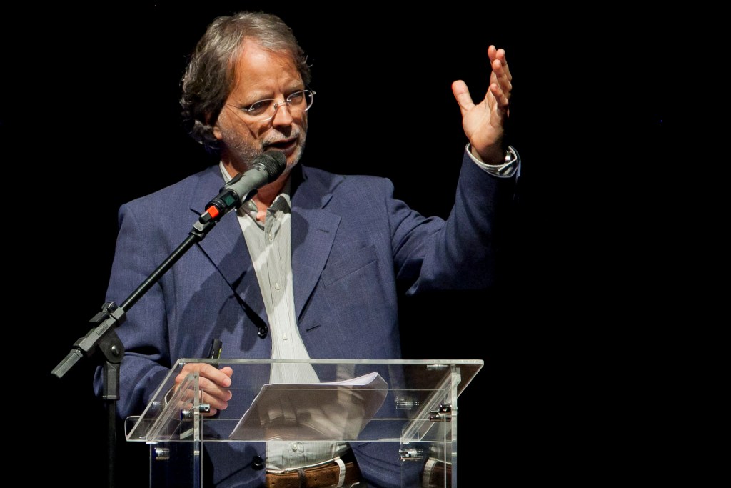 Mia Couto speaking passionately at an event, gesturing with one hand while holding a microphone, behind a transparent podium.