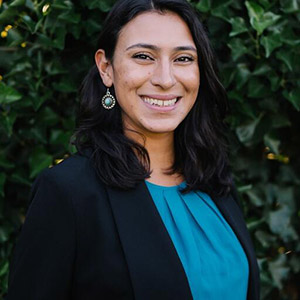 A woman smiling while wearing a black blazer and a blue top, with earrings, in front of a green leafy background.