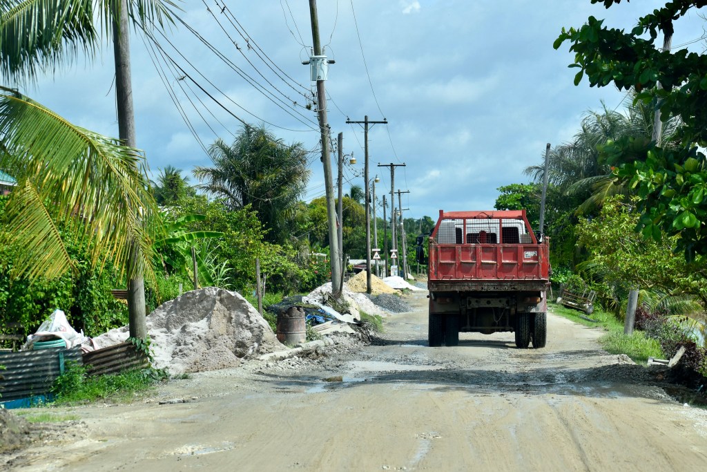 A red truck driving on a dirt road lined with palm trees and power poles, surrounded by piles of sand and building materials.