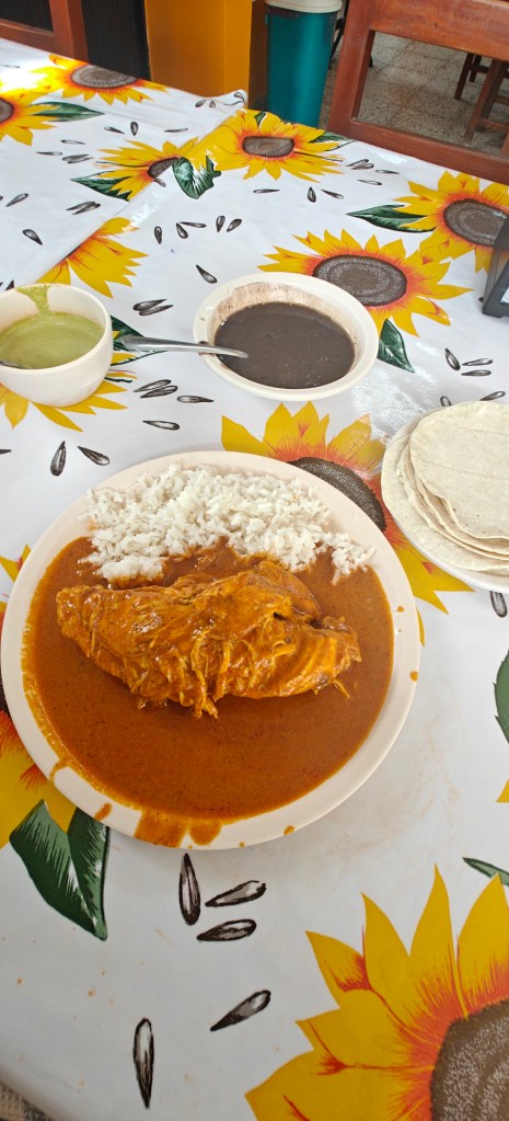 A plate of rice served with a piece of chicken in a rich, orange sauce, accompanied by bowls of green and black sauces and a stack of tortillas on a colorful sunflower-patterned tablecloth.