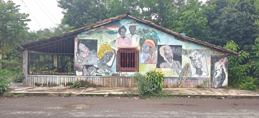 Colorful mural depicting portraits of women on the exterior of a house, surrounded by greenery.