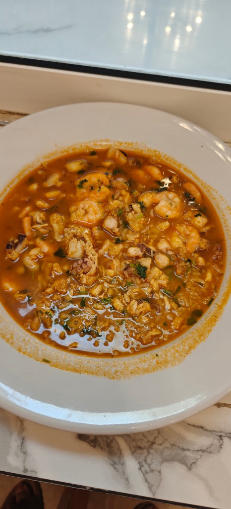 A bowl of hearty seafood soup with shrimp and chunks of fish, garnished with herbs, served on a marble table.
