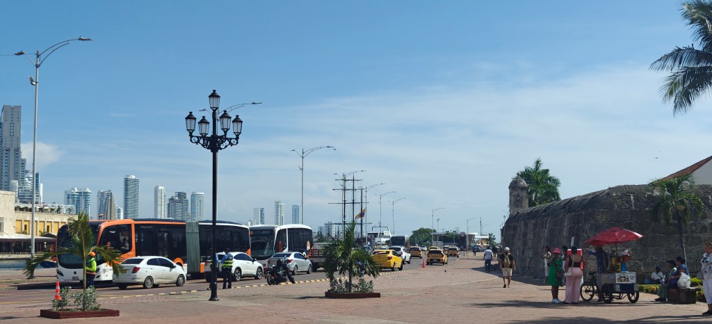 A busy street scene in a coastal city with buses, cars, and people, featuring palm trees, a historic wall, and street vendors under bright blue skies.