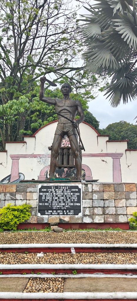 Statue of 'El Yanga,' a historical figure, holding a sword, surrounded by greenery in a public space, with a plaque detailing his significance.
