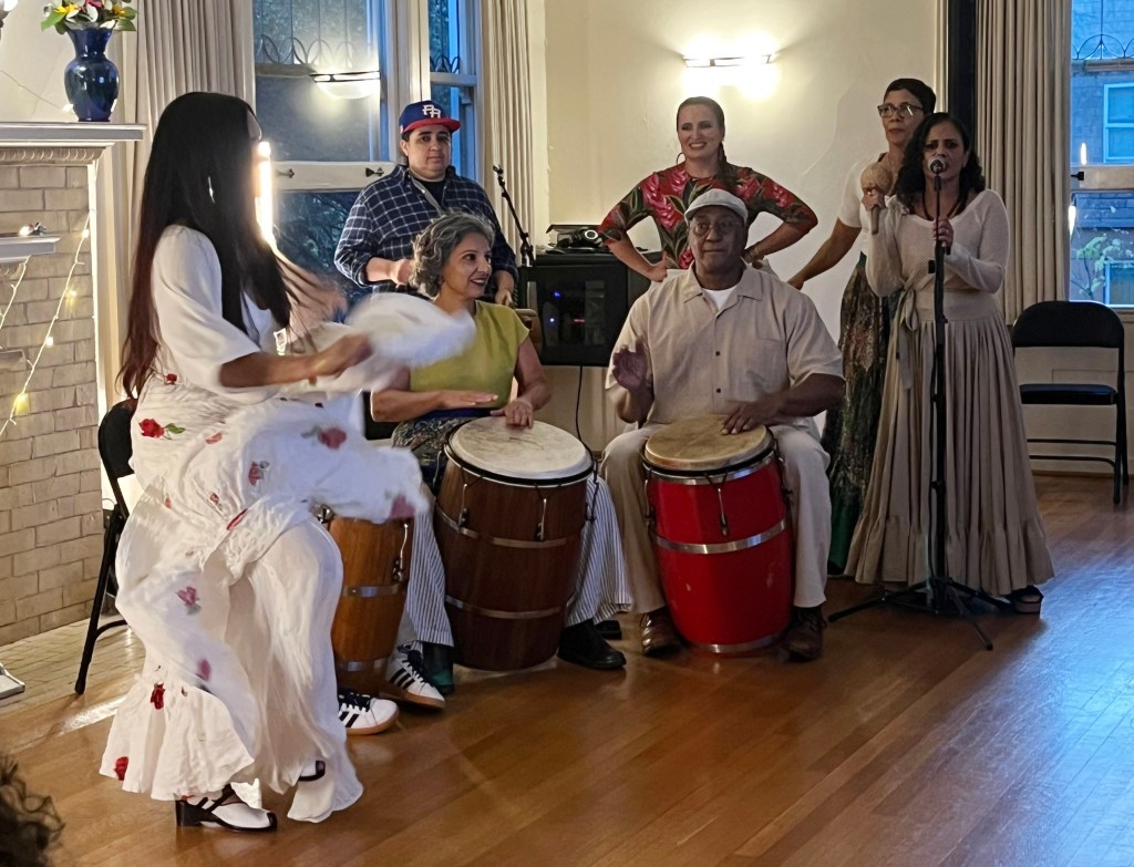 A group of musicians performing in a cozy indoor setting. One woman is dancing in a flowing white dress, while others are playing drums and singing. A man stands in the background, watching the performance, and additional performers are singing on stage.