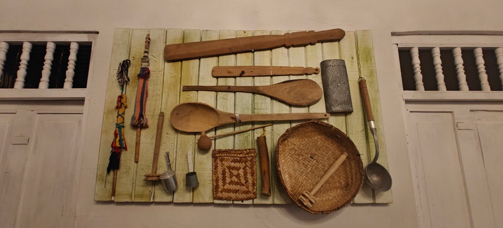 A collection of traditional kitchen utensils displayed on a wooden board, including wooden spoons, a grater, a woven basket, and decorative woven items.