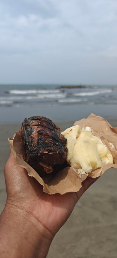 A hand holding fried fish and boiled yucca in front of waves on a beach.