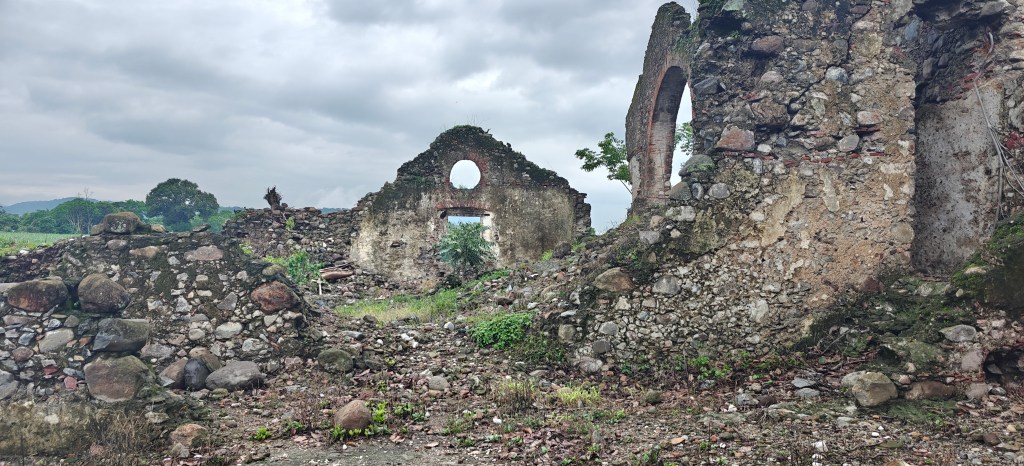 Ruins of a stone structure with broken walls and arched openings, surrounded by greenery and rocky terrain under a cloudy sky.
