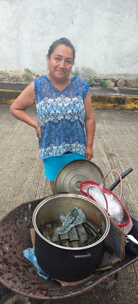 A woman standing behind a pot over a fire outside on a sidewalk.