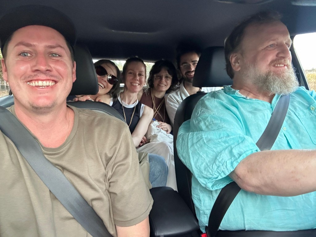 A group of six people smiling while sitting in a car, with one person driving. The atmosphere is cheerful and relaxed.