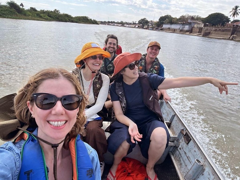 A group of five people on a boat, smiling and enjoying a day on the water. The woman in front wears sunglasses and a life jacket. The others are dressed casually with various hats, surrounded by scenic riverbanks.