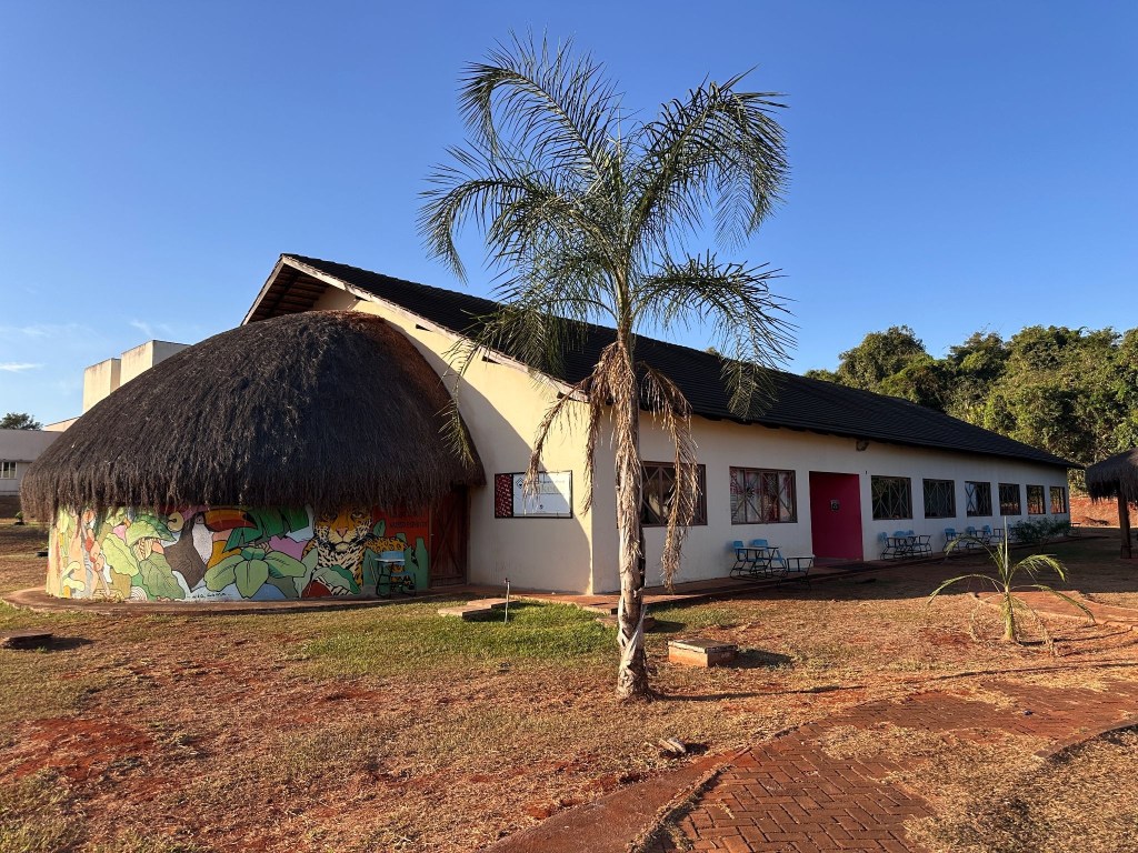 A building with a thatched roof on the left and a white structure on the right, surrounded by grass and trees under a clear blue sky.