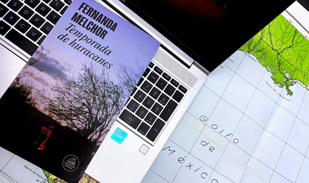 A top-down view of a laptop keyboard with a book titled 'Temporada de huracanes' by Fernanda Melchor resting on it, alongside a folded map showing the Gulf of Mexico.