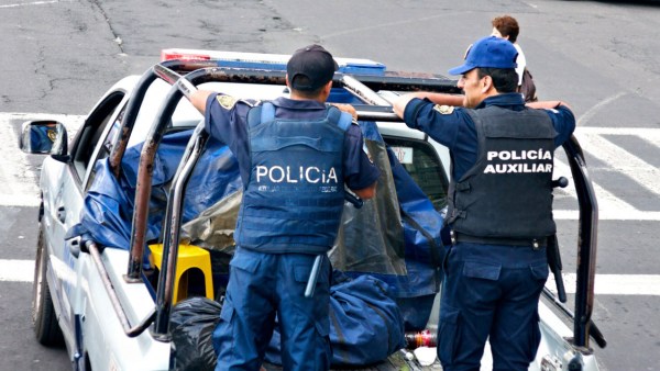 Mexican police in blue uniforms patrol a street standing in the back of a white pickup truck.
