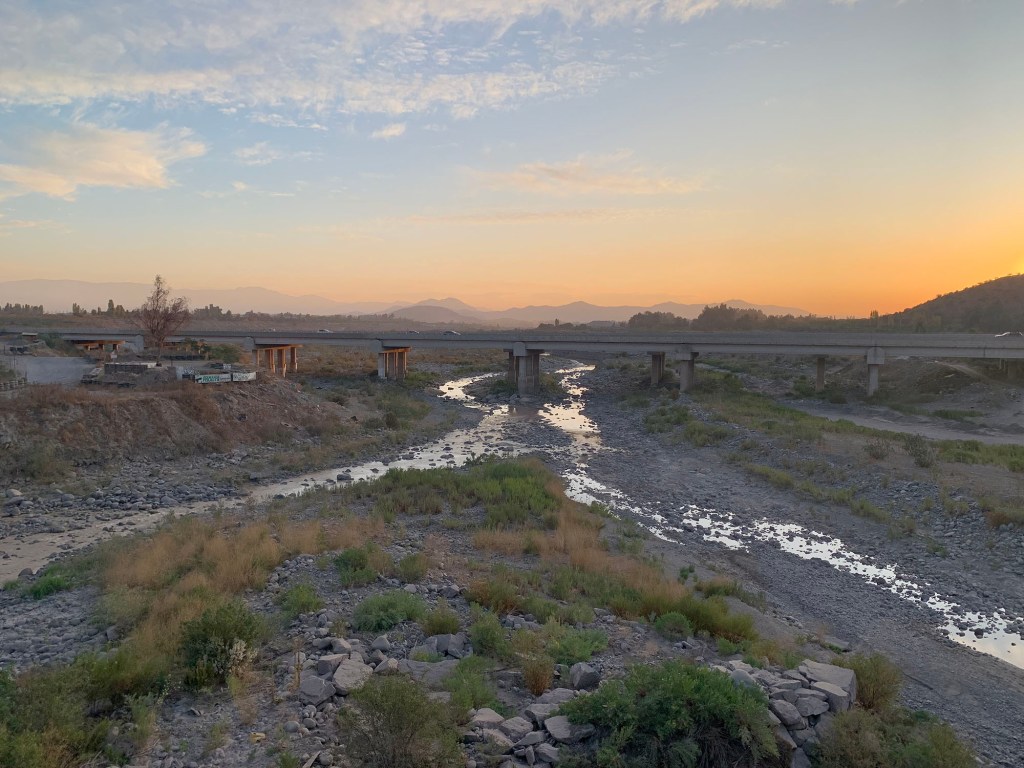 A serene landscape featuring a riverbed with a shallow stream flowing through it, surrounded by rocky terrain and sparse vegetation. In the background, a bridge spans the river, under a colorful sunset sky with distant mountains.