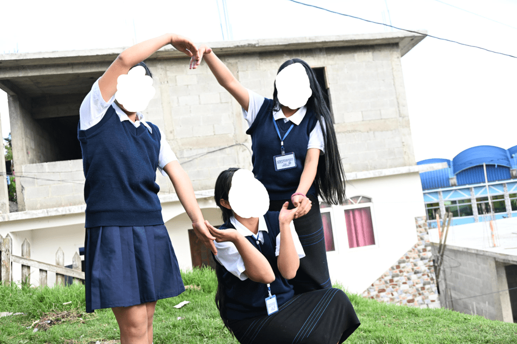 Three girls in school uniforms stand with their arms formed into a heart shape.