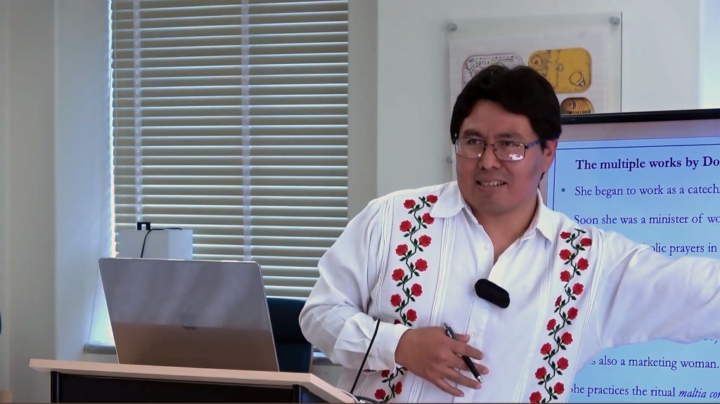 A man in glasses and a white shirt with embroidered red flowers speaks while pointing to an onscreen presentation.
