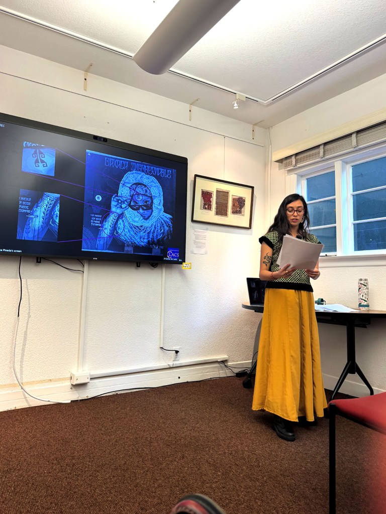 A speaker in a yellow skirt reads from papers in front of a presentation in a small conference room.