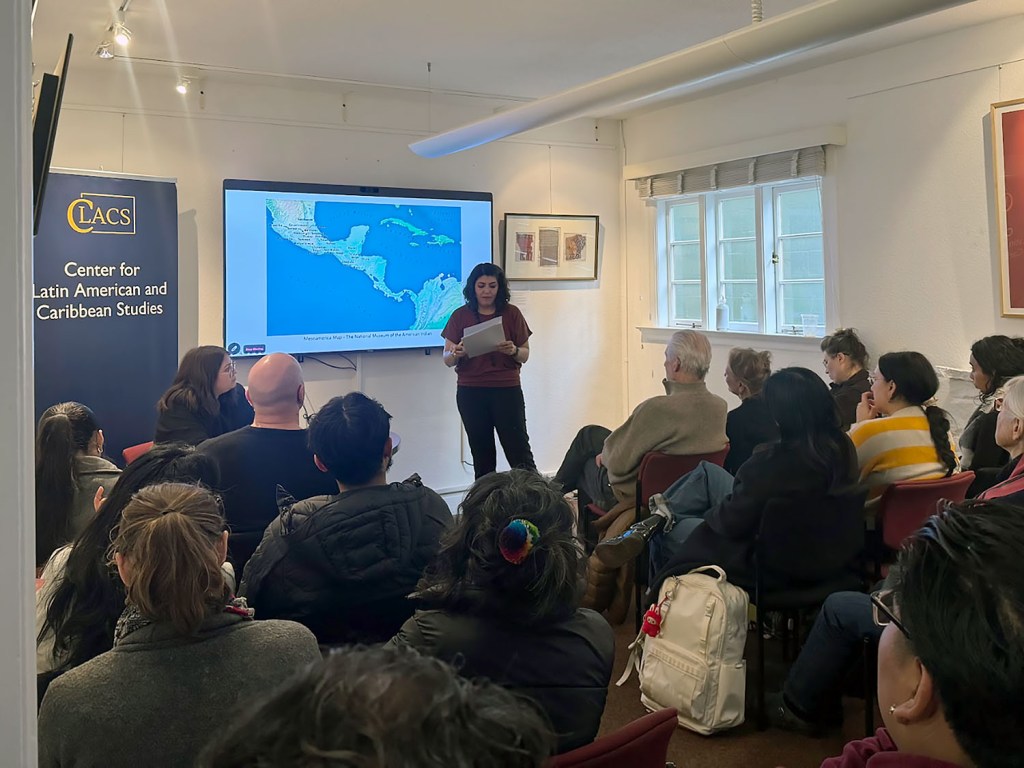 A speaker stands reading from papers in front of a map of Central America.