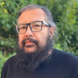A man with a beard and glasses, wearing a black sweater, standing outdoors with greenery in the background.
